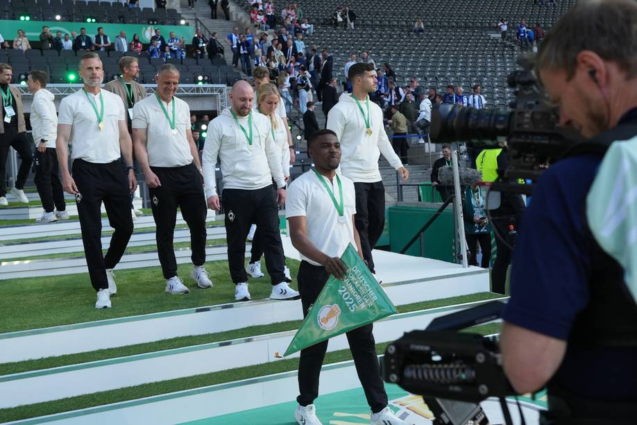 Cédric Makiadi hält im Vordergrund den Wimpel für den Sieg im DFB-Pokal 2025 in der Hand. Links hinter im folgt ihm sein Trainerteam im Olympiastadion.