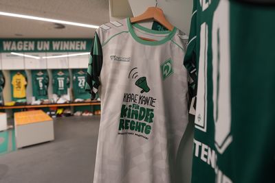 In einem Umkleideraum hängt ein grün-weißes Sporttrikot mit einer Megafon-Grafik und einem Text, der für Kinderrechte wirbt.