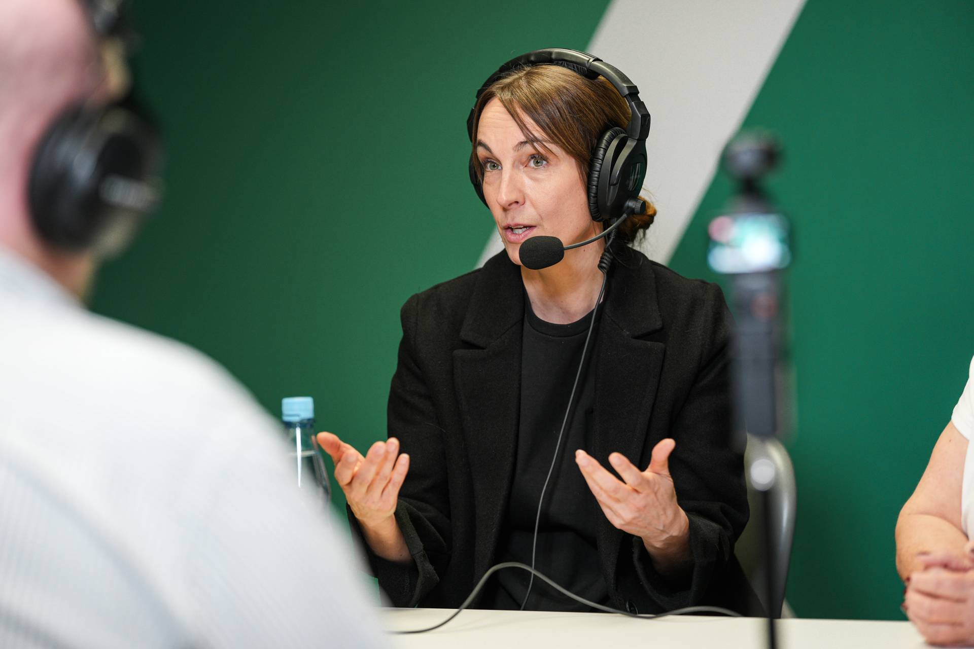 Eine Frau mit Kopfhörern spricht während einer Podcast-Aufnahme in einem Studio mit grünem Hintergrund in ein Mikrofon.