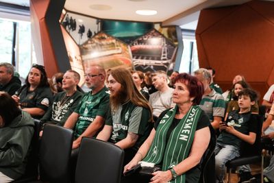 Eine Gruppe von Fußballfans sitzt in einem Innenraum und schaut aufmerksam nach vorne. Viele tragen grüne Fanartikel und Trikots, einige haben Schals um den Hals. Im Vordergrund sitzt eine ältere Frau mit kurzem roten Haar und grünem Schal, daneben ein Mann mit langem Haar und Bart im Trikot. Im Hintergrund sind weitere Zuschauer, darunter auch ein Kind, sowie Wandbilder eines Stadions zu sehen.