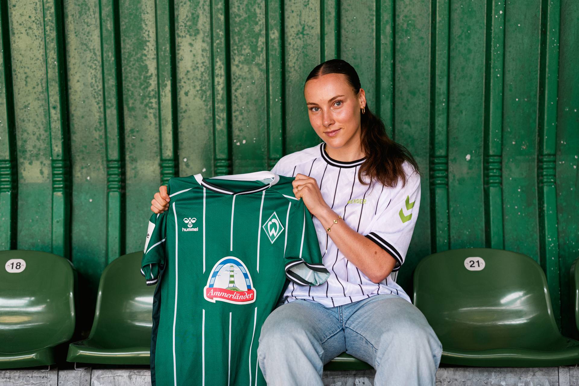 Felicia Sträßer sitzt vor einer grünen Wand und hält ein Werder-Trikot hoch.