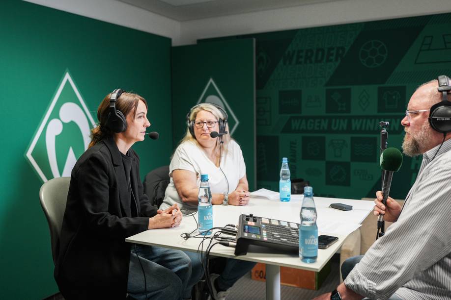 Drei Personen mit Kopfhörern sitzen in einem Tonstudio mit Mikrofonen um einen Tisch herum.