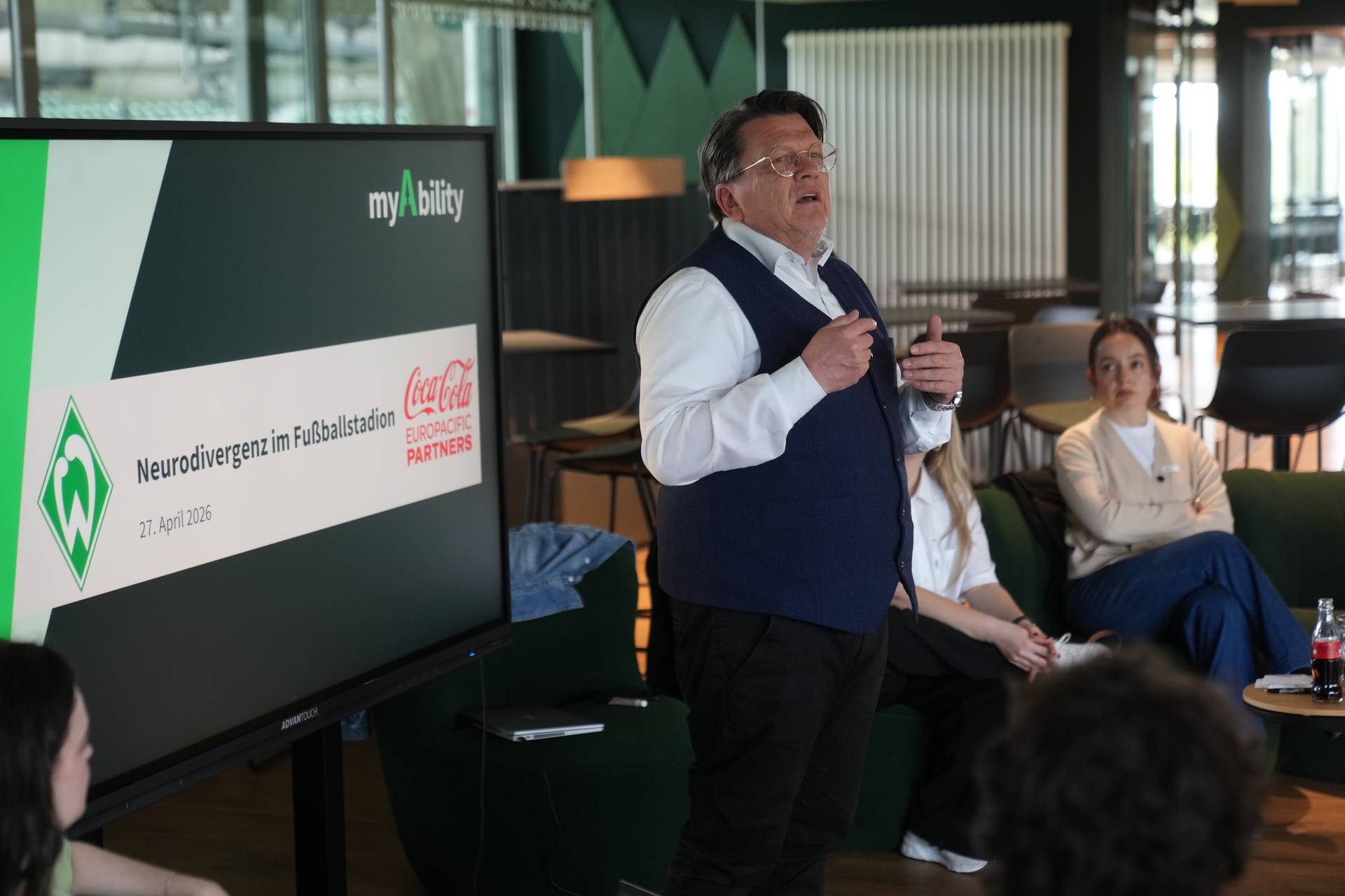 Ein Mann hält vor einem Publikum, das in einem modernen Raum sitzt, einen Vortrag über Neurodiversität im Fußball, wobei das Thema auf einer Leinwand im Hintergrund angezeigt wird.