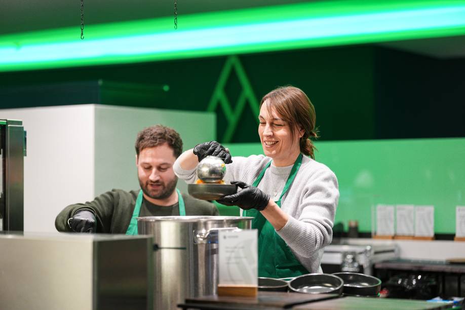 Zwei Personen, die Schürzen und Handschuhe tragen, servieren Speisen aus einem großen Topf in einer Küche mit hellgrüner Beleuchtung.