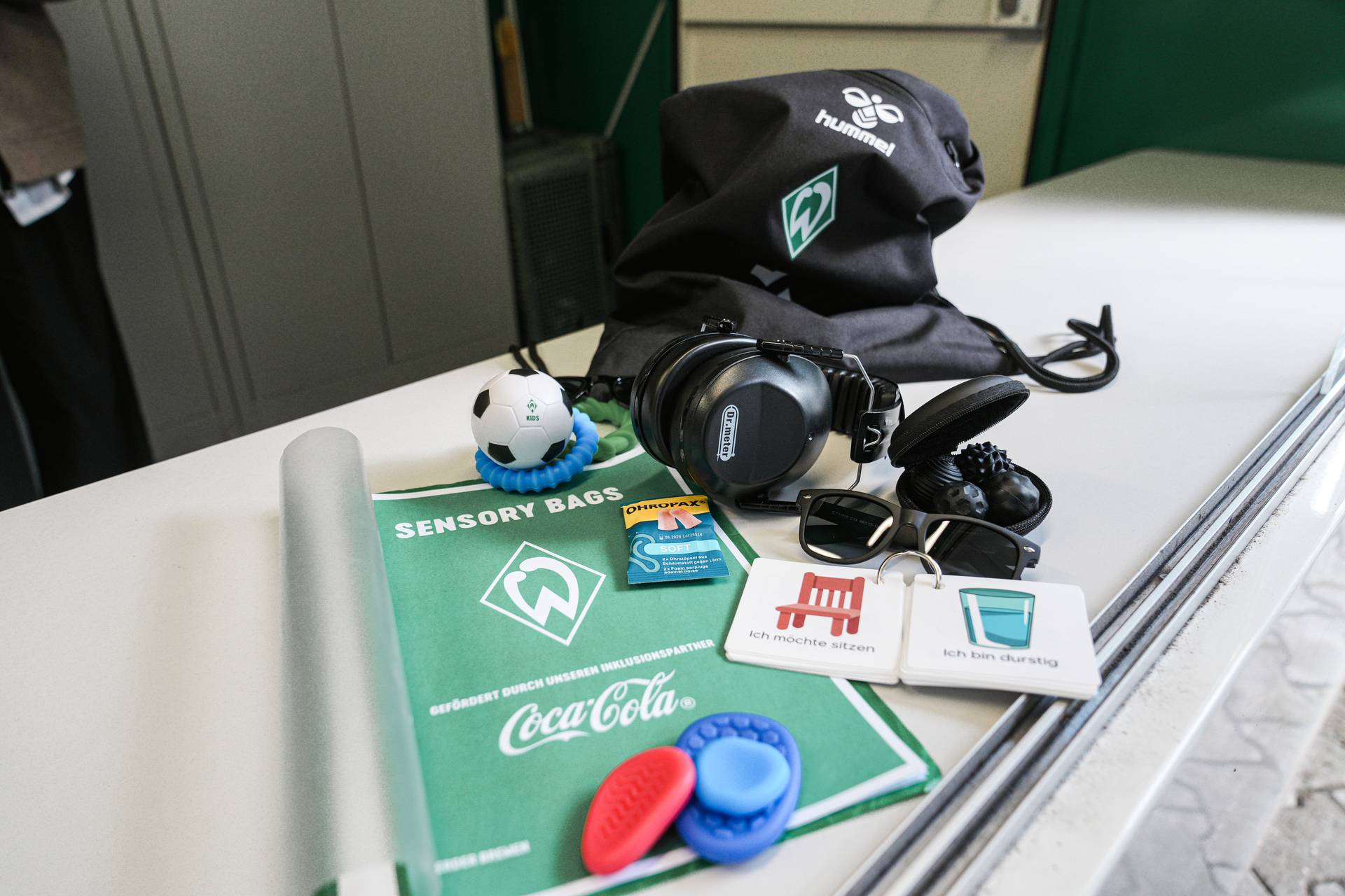 Inhalt des Sensoriktäschchens: Kopfhörer, Stressball, Ohrstöpsel, Sonnenbrille, Sensorikkarten, grüne Matte und ein schwarzer Beutel mit Kordelzug auf einem Tisch.