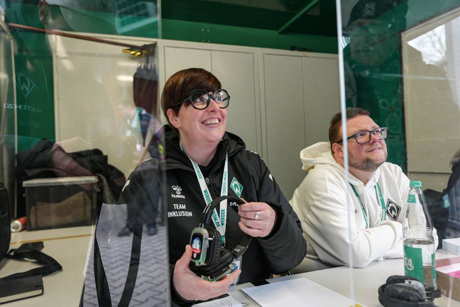 Zwei Menschen mit Jacken an und Lanyards um den Hals sitzen hinter einer Glasscheibe, lachen, schauen nach vorne und geben Kopfhörer nach vorne heraus.