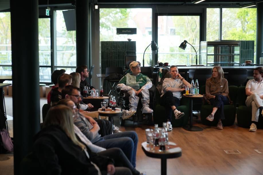 Menschen sitzen in einem Halbkreis in einer ungezwungenen Umgebung in einem Innenraum und unterhalten sich. Auf dem Tisch stehen Getränke und Flaschen.