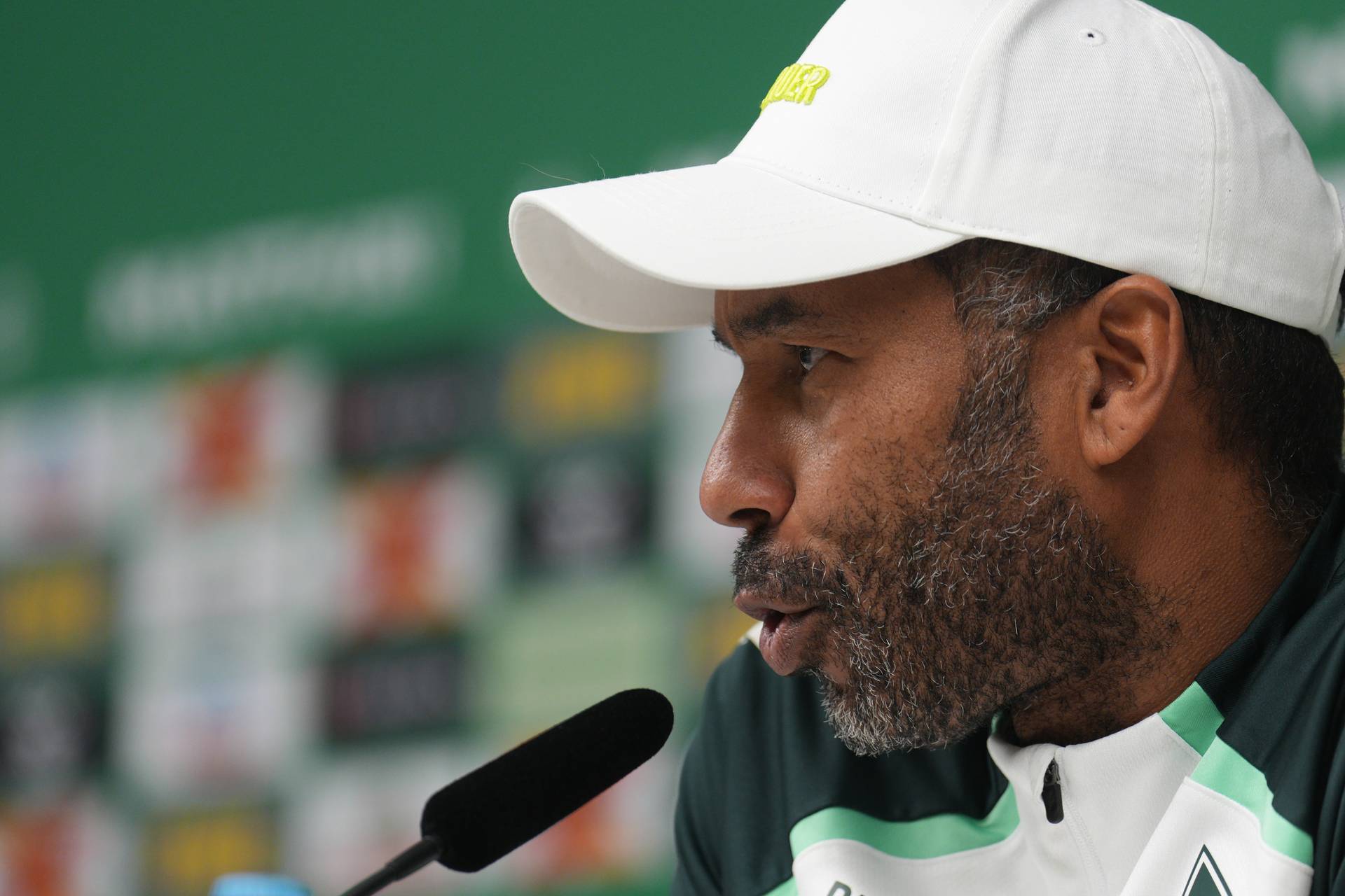 Daniel Thioune mit weißer Werder-Kappe auf dem Podium bei der Pressekonferenz seitlich