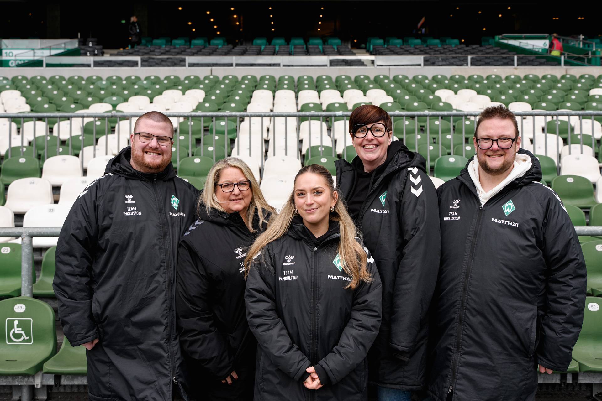 Fünf Personen in einheitlichen schwarzen Jacken stehen vor grün-weißen Stadion-Sitzen und lächeln in die Kamera.