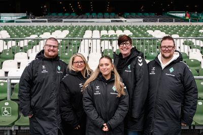 Fünf Personen in einheitlichen schwarzen Jacken stehen vor grün-weißen Stadion-Sitzen und lächeln in die Kamera.