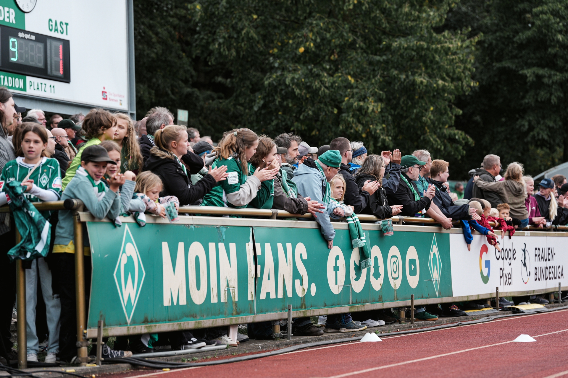 Fans in grüner Kleidung versammeln sich begeistert an einer Brüstung bei einem Fußballspiel, im Hintergrund sind Schilder und Werbeplakate zu sehen.
