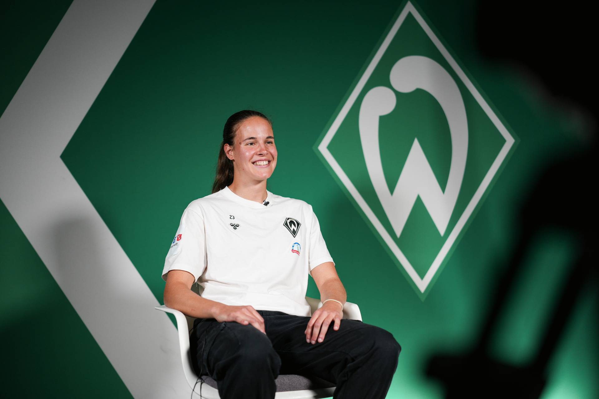 Hanna Nemeth sitzt lächelnd vor einem grünen Hintergrund mit einem Logo, das dem von Werder Bremen ähnelt, und trägt ein weißes Shirt mit Logo.
