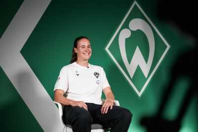 Hanna Nemeth sitzt lächelnd vor einem grünen Hintergrund mit einem Logo, das dem von Werder Bremen ähnelt, und trägt ein weißes Shirt mit Logo.
