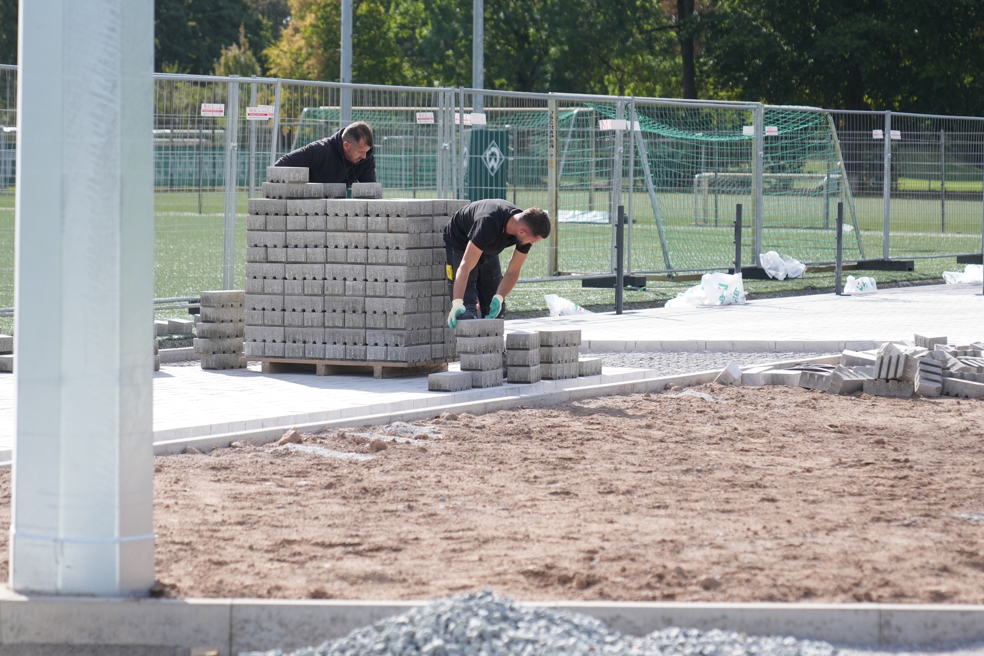 An einem sonnigen Tag legen zwei Arbeiter neben einem umzäunten Sportplatz Pflastersteine bereit, um den Pflasterbau vorzubereiten.