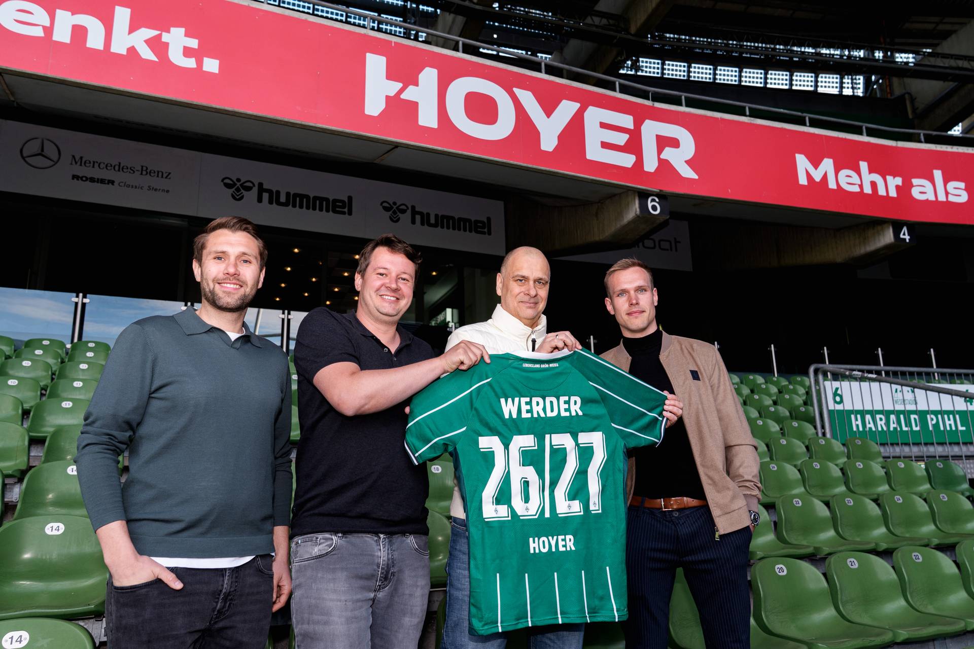 Vier Männer stehen in einem Stadion, lächeln und halten ein grünes Werder-Trikot mit den Aufschriften „26/27“ und „HOYER" auf dem Rücken in den Händen.