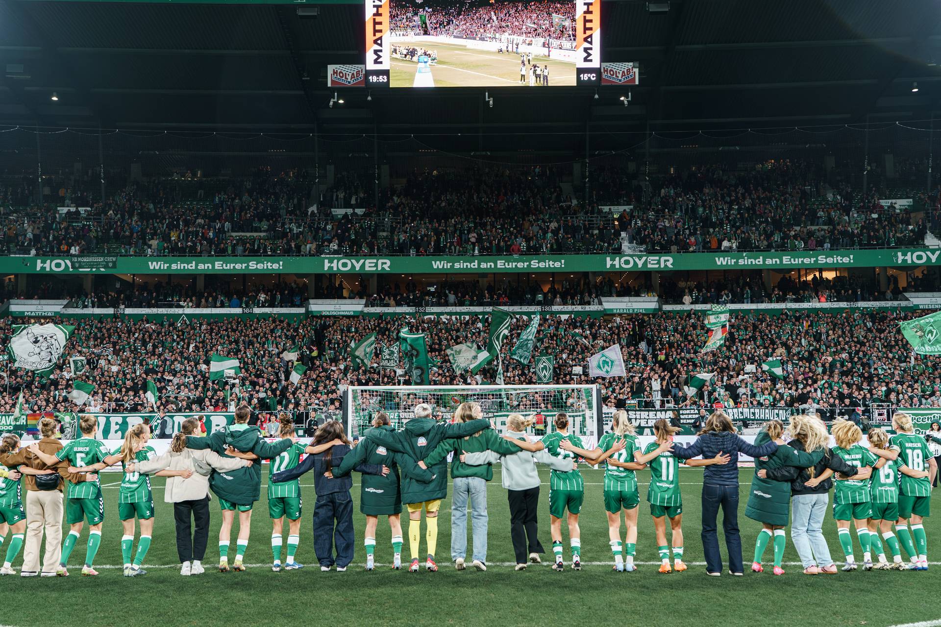 Die WERDERFRAUEN auf dem Spielfeld, Arm in Arm, im Hintergrund eine ausgelassene Zuschauermenge, die unter den Stadionlichtern grüne Fahnen schwenkt.