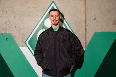 Marco Friedl in einer Bomberjacke vor einer Wand mit Werder-Raute.
