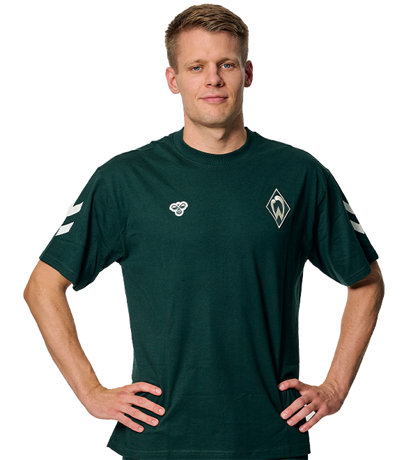 Person in a dark green sports shirt with a logo on the chest, standing with hands on hips against a white background.