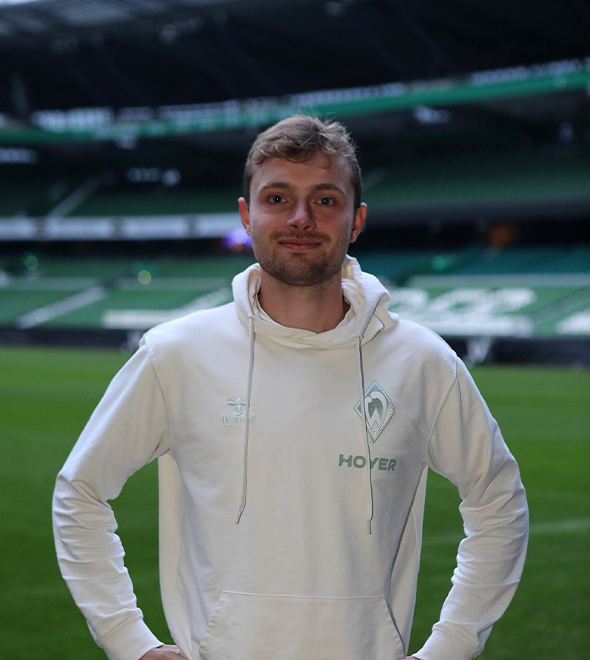 Leander Wasmus steht in einem weißen Kapuzenpulli auf einem Fußballfeld, im Hintergrund sind leere grüne Stadionplätze zu sehen.