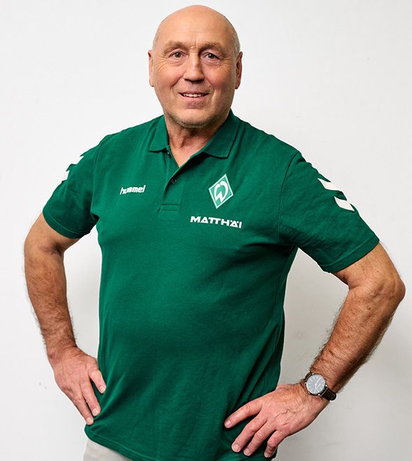 Ein lächelnder Mann in einem grünen Sport-Polohemd mit Logos steht selbstbewusst mit den Händen in den Hüften vor einem schlichten weißen Hintergrund.