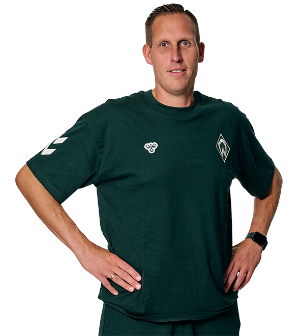 Person in a dark green sports shirt with logos, standing with hands on hips, against a white background.