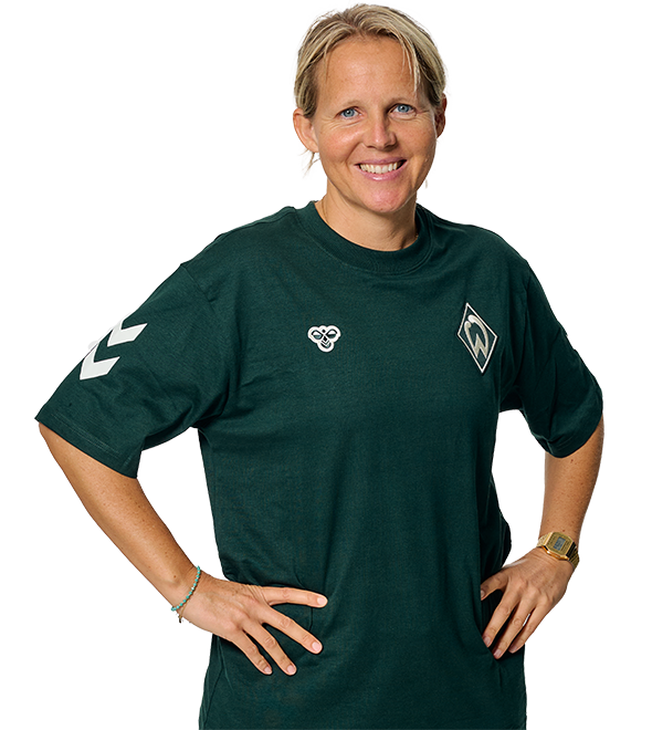 Person smiling, wearing a dark green sports jersey with logos, hands on hips, short blonde hair, and a watch on the left wrist.