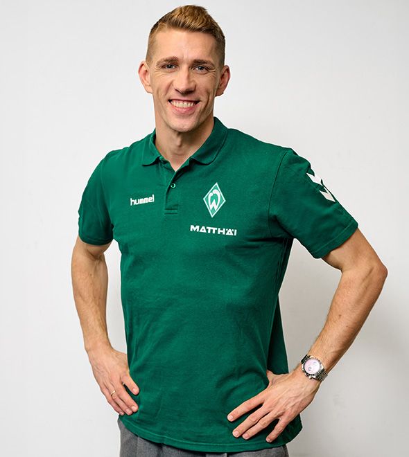 Ein lächelnder Mann in einem grünen Sport-Polohemd mit Logos steht selbstbewusst mit den Händen in den Hüften vor einem schlichten weißen Hintergrund.