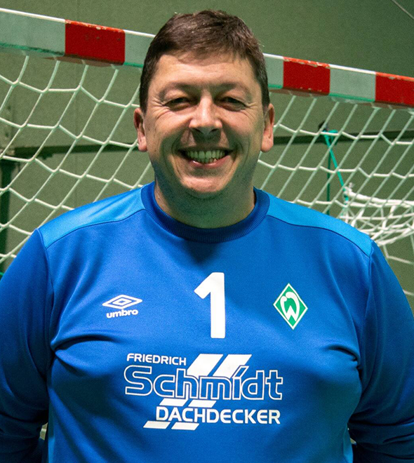 Ein lächelnder Mann in einem blauen Sporttrikot steht vor einem Fußballtor in einer Halle.