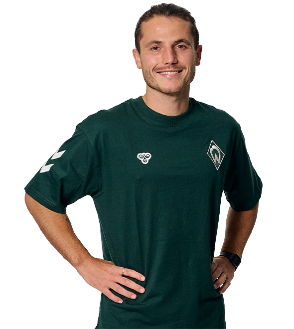 Smiling person wearing a green sports shirt with logos, standing with hands on hips against a white background.