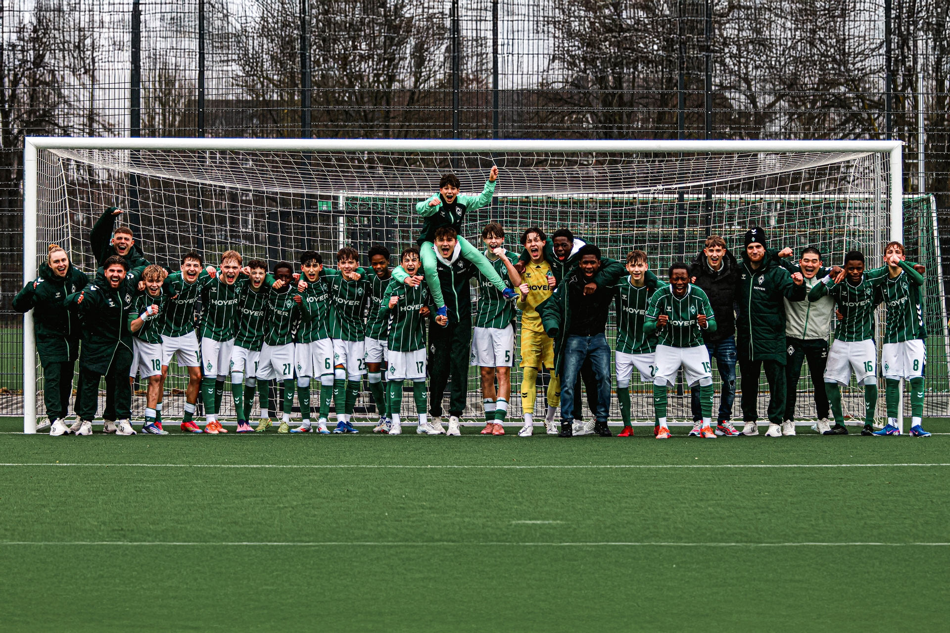Die Trainer*innen und Spieler stehen in einer Reihe in einem Fußballtor und jubeln.