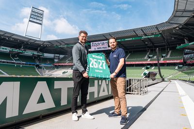 Zwei Männer, die in einem leeren Fußballstadion ein Trikot von Werder Bremen mit der Nummer 26/27 und dem Namen TOPRAN in den Händen halten.