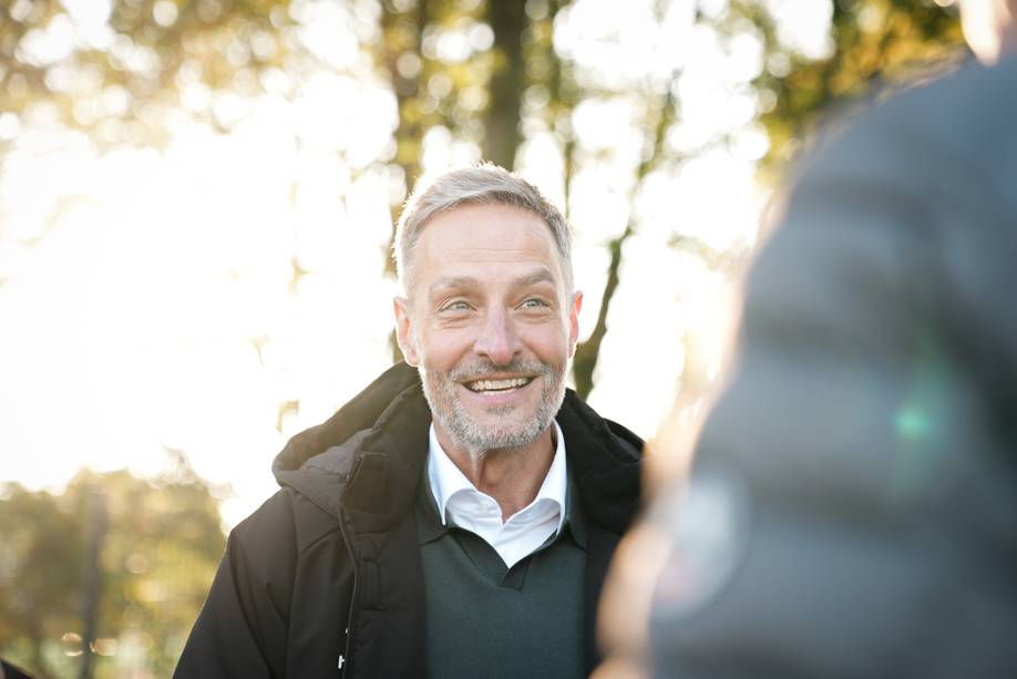 Ein lächelnder Mann mit kurzen grauen Haaren steht im Sonnenlicht im Freien, trägt eine dunkle Jacke und einen Pullover, im Hintergrund sind verschwommene Bäume zu sehen.