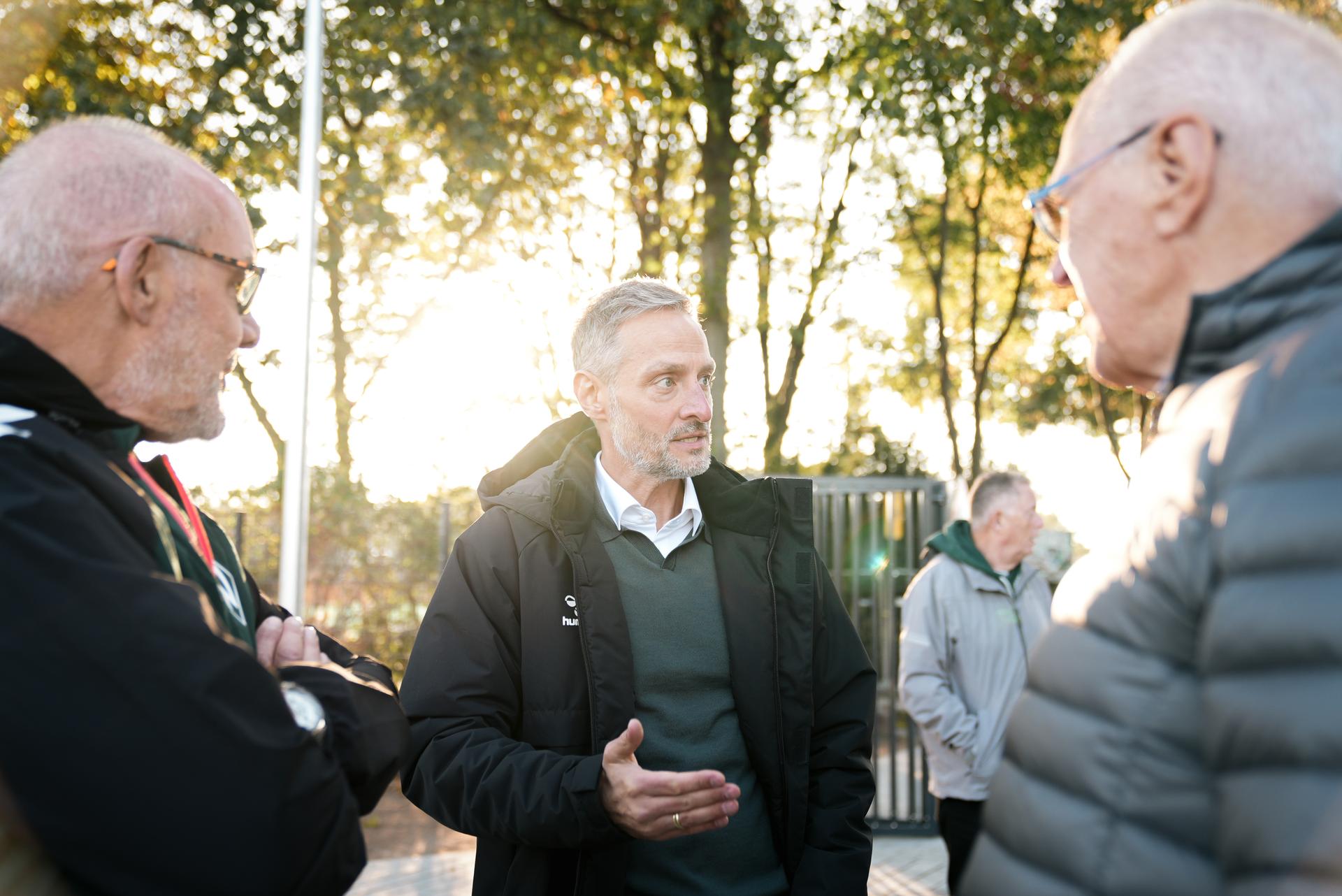 Drei Männer unterhalten sich im Freien, im Hintergrund sind Bäume und Sonnenlicht zu sehen. Ein Mann gestikuliert beim Sprechen.