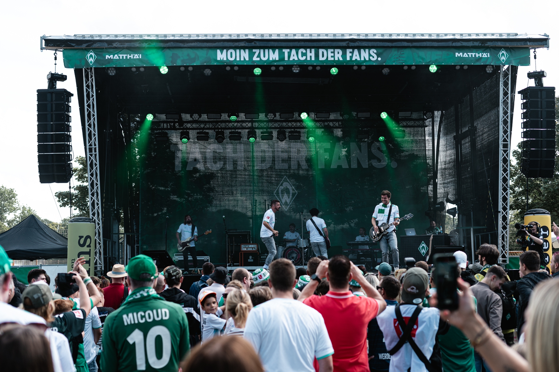 Open-Air-Konzert mit einer Band auf der Bühne, umgeben von Fans in grün-weißen Sporttrikots. Auf dem Banner über der Bühne steht „Moin zum Tach der Fans“.