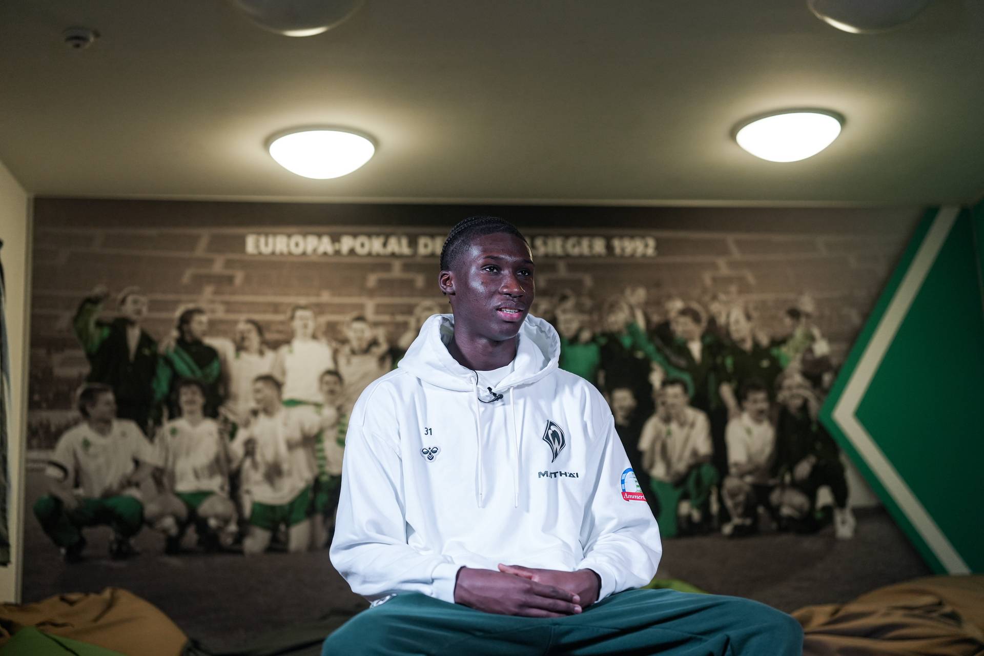 Karim Coulibaly sitzt in einem weißen Pullover vor einer Wand, auf dem das Siegerfoto der Europa-Pokal der Pokalsieger 1992 zu sehen ist.