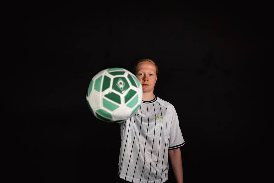 Thea Farwick, die ein weißes Fußballtrikot mit Nadelstreifen trägt, hält vor einem schwarzen Hintergrund einen grün-weißen Fußball in Richtung Kamera.
