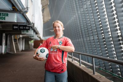 Elena Dhont in einem roten Trikot von Werder Bremen, einen Fußball in der Hand und lächelnd, steht in einem Stadiongang.