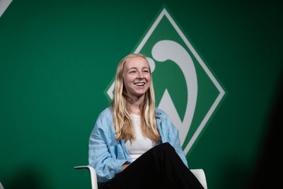 Caro Siems sitzt in einem blauen Hemd auf einem Stuhl vor einem grünen Hintergrund mit einem weißen Logo.