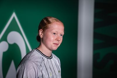 Thea Farwick trägt ein Sporttrikot und steht vor einem grün-weißen Hintergrund mit einem stilisierten Logo.