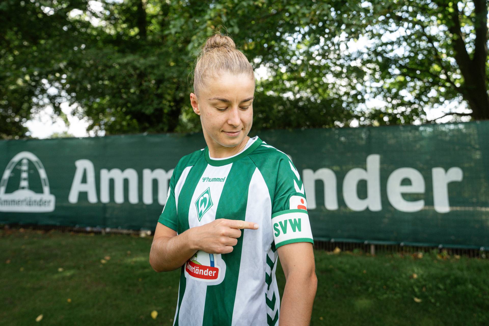 Michelle Ulbrich, die ein grün-weißes Trikot trägt, zeigt auf eine Kapitänsbinde mit der Aufschrift „SVW“, während sie neben einem Zaun mit einem Logo steht.