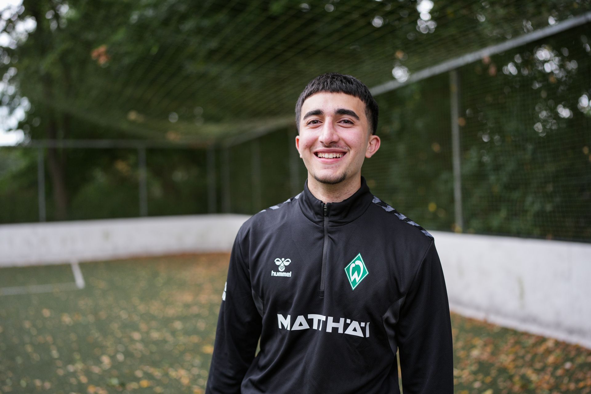 Hasan Hourou steht auf einem Bolzplatz und lächelt in die Kamera.