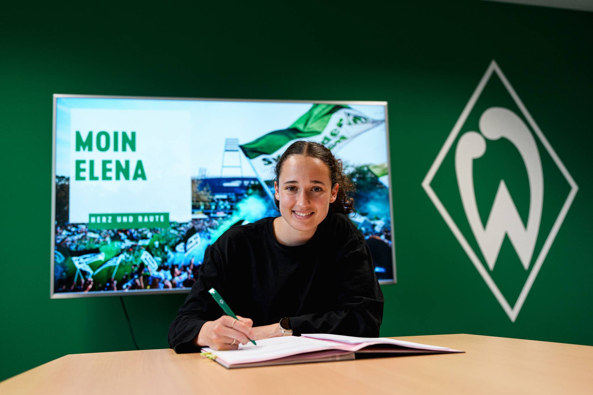 Elena Mühlemann unterschreibt an einem Tisch ein Dokument, hinter ihr ist ein grüner Hintergrund mit der Aufschrift „Moin Elena“ zu sehen, und an der Wand hängt das Logo einer Sportmannschaft.