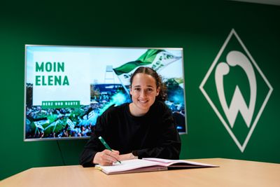 Elena Mühlemann unterschreibt an einem Tisch ein Dokument, hinter ihr ist ein grüner Hintergrund mit der Aufschrift „Moin Elena“ zu sehen, und an der Wand hängt das Logo einer Sportmannschaft.