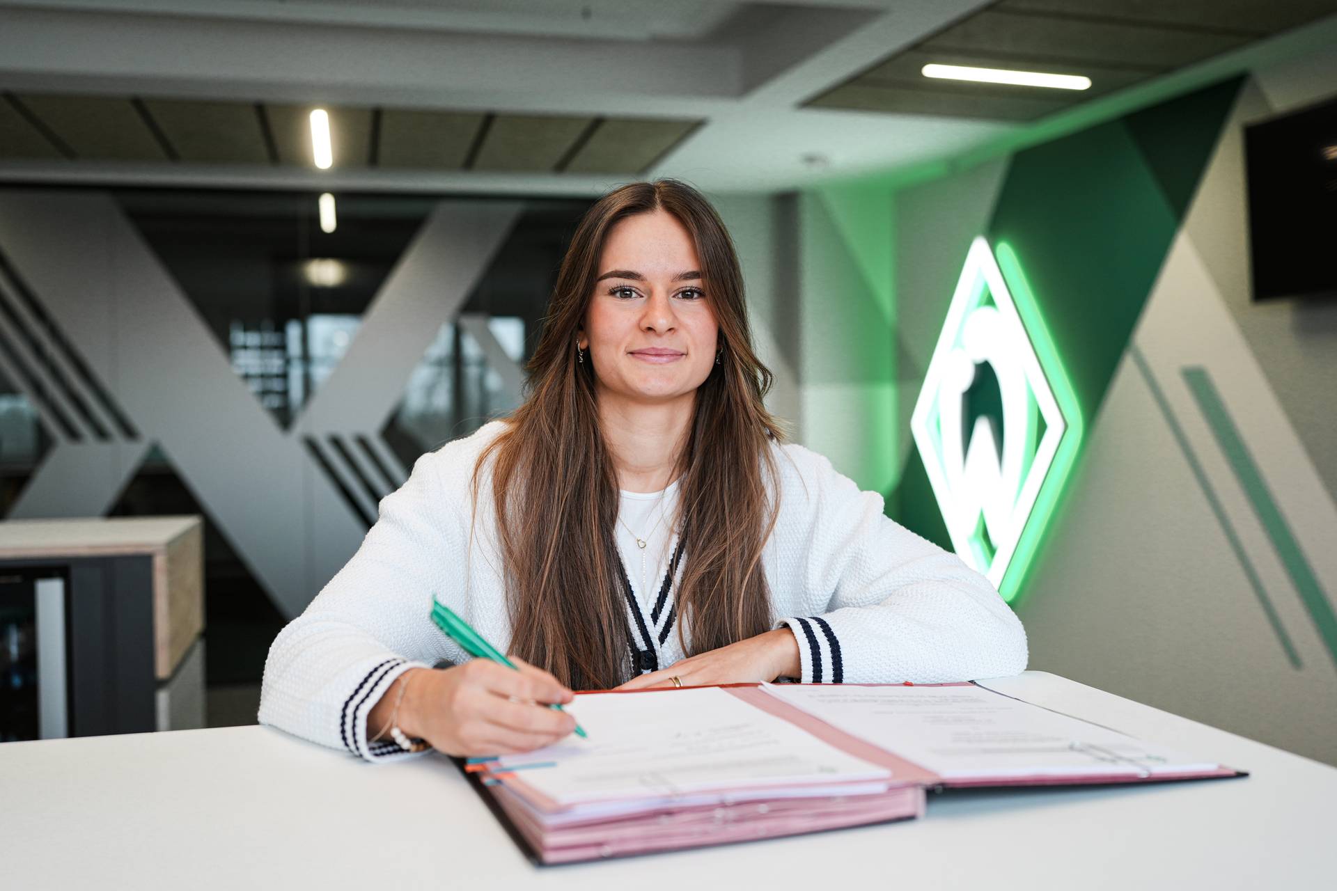 Tuana Mahmoud unterschreibt ihren Vertrag und lächelt dabei in die Kamera. Im Hintergrund ist eine leuchtende Werder-Raute zu sehen.