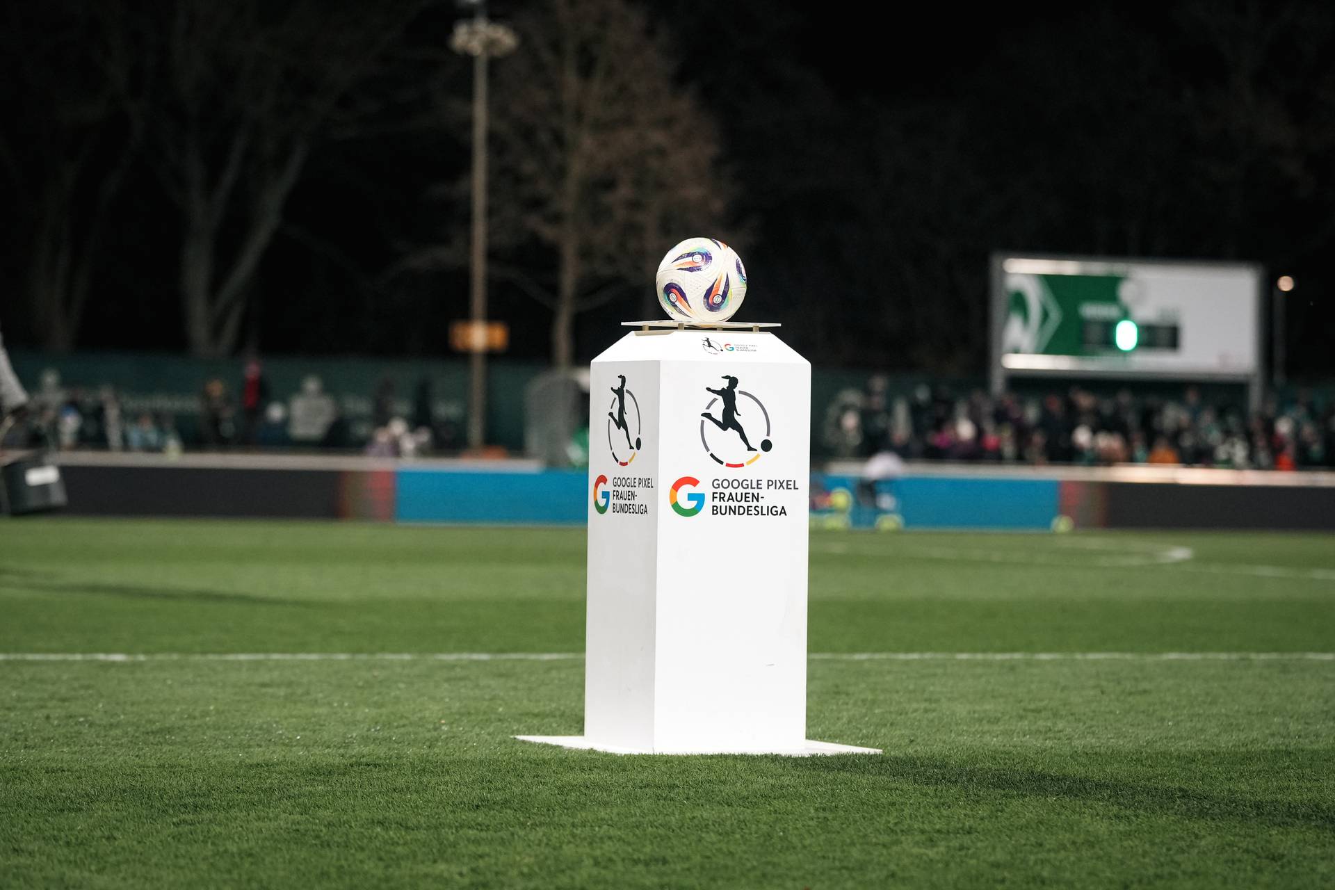 Ein Fußball liegt auf einem weißen Sockel mit den Logos der Google Pixel Frauen-Bundesliga, vor dem Hintergrund eines grünen Spielfelds mit verschwommenen Zuschauern.