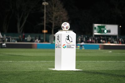 Ein Fußball liegt auf einem weißen Sockel mit den Logos der Google Pixel Frauen-Bundesliga, vor dem Hintergrund eines grünen Spielfelds mit verschwommenen Zuschauern.