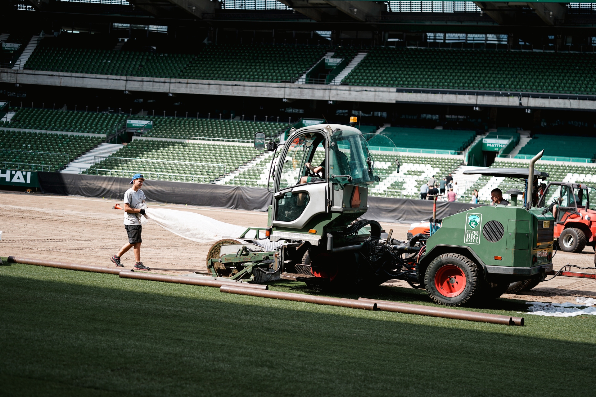 Arbeiter bedienen Maschinen auf einem Sportplatz und bereiten den Boden vor. Die Tribünen im Hintergrund sind leer.