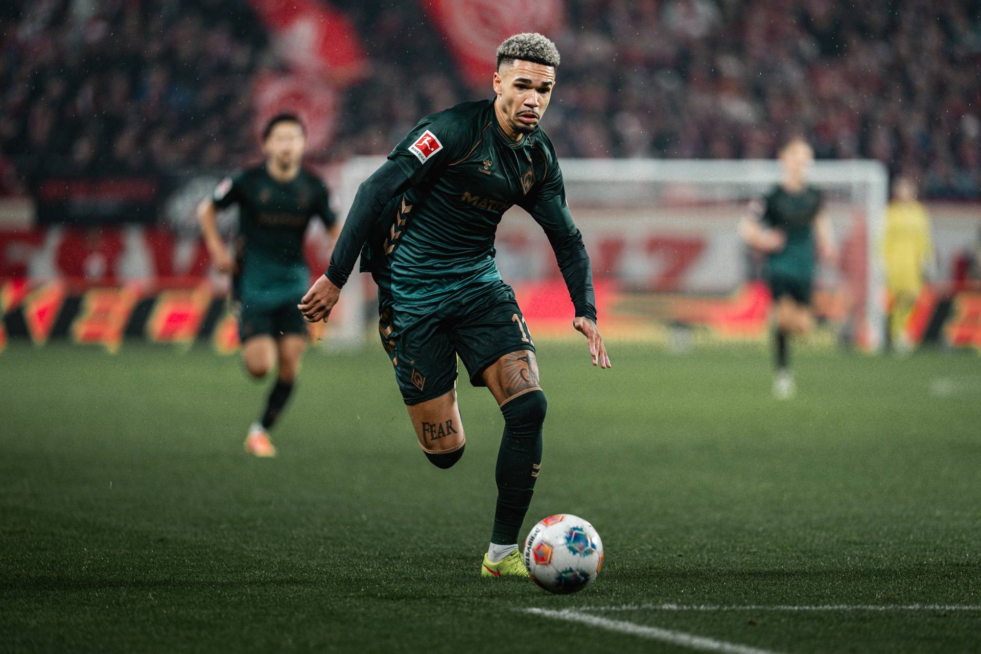 Justin Njinmah läuft im dunkelgrünen Third-Trikot mit dem Ball am Fuß im Stadion.