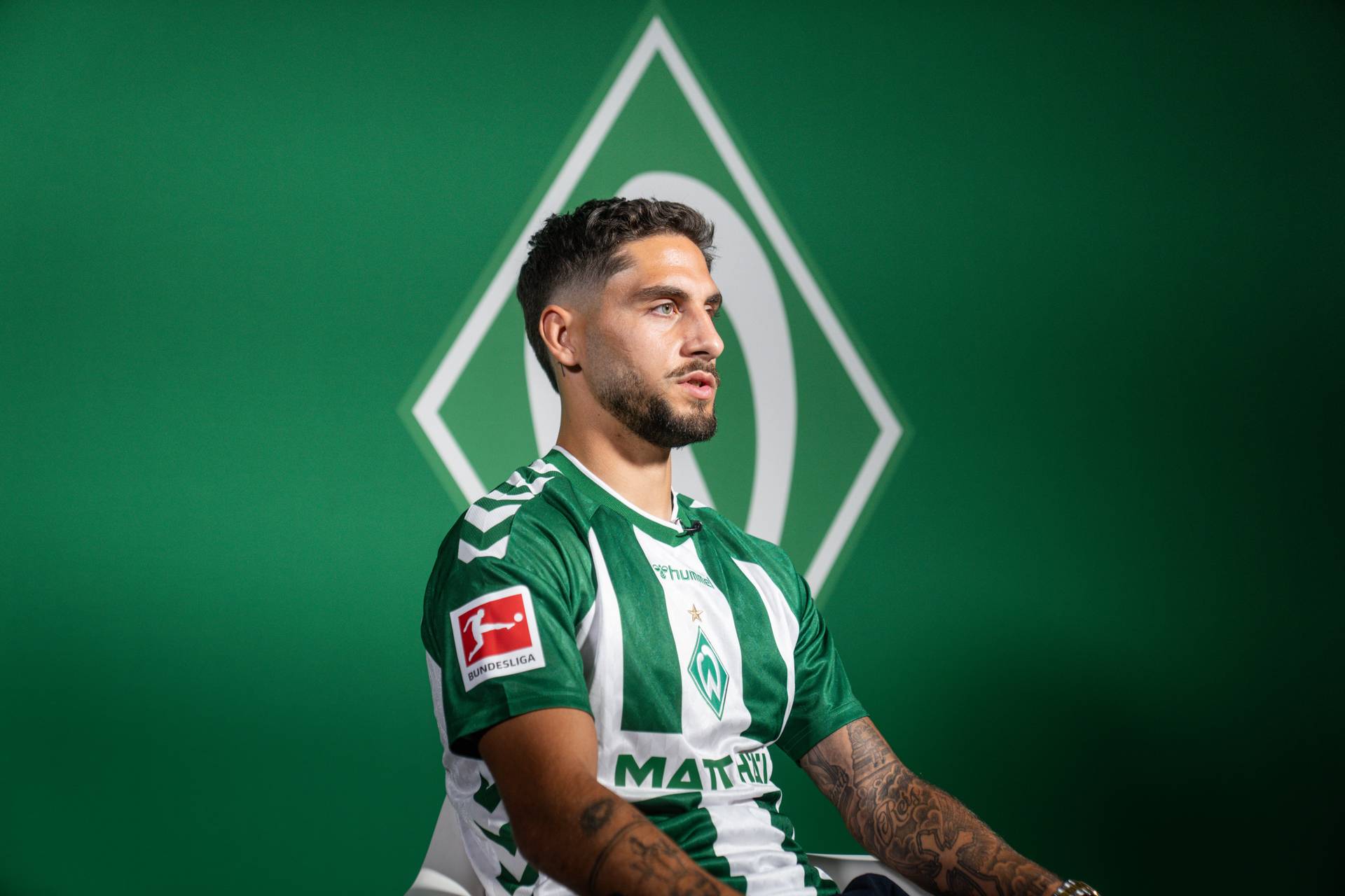 Ludovit Reis sitzt im Heimtrikot vor einer grünen Wand mit großer Werder-Raute.