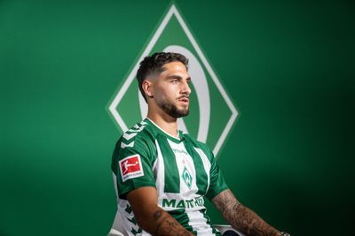 Ludovit Reis sitzt im Heimtrikot vor einer grünen Wand mit großer Werder-Raute.